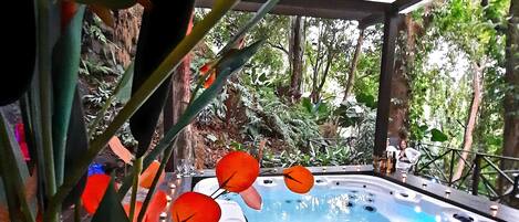 A hot tub surrounded by greenery, with a wooden deck and a pergola overhead.