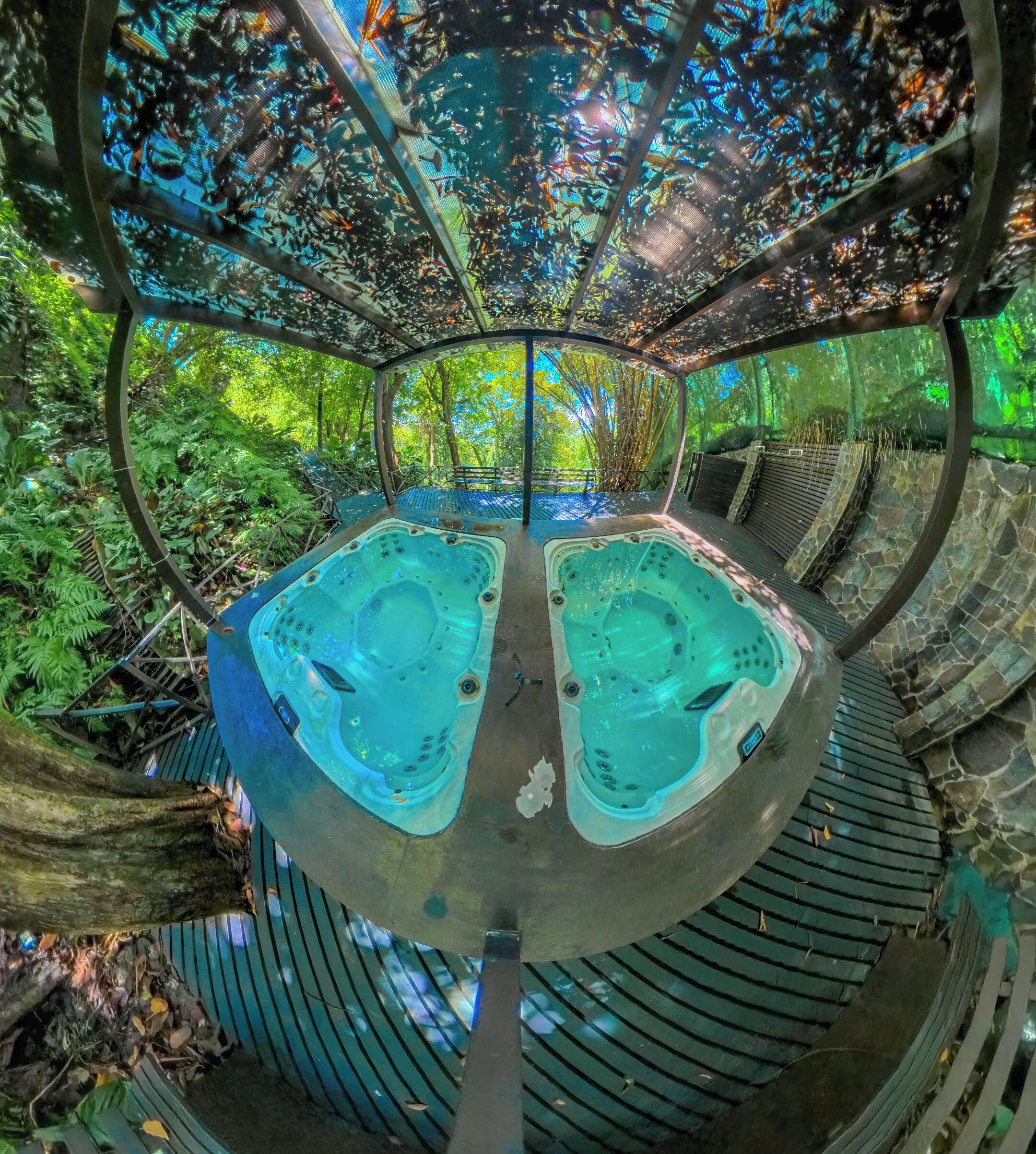 A circular hot tub structure with two hot tubs, surrounded by a wooden deck and lush greenery.