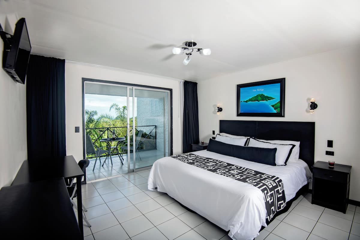 Una habitación de hotel moderna con una cama grande, un televisor de pantalla plana montado en la pared, un ventilador de techo y un balcón con puerta de vidrio.