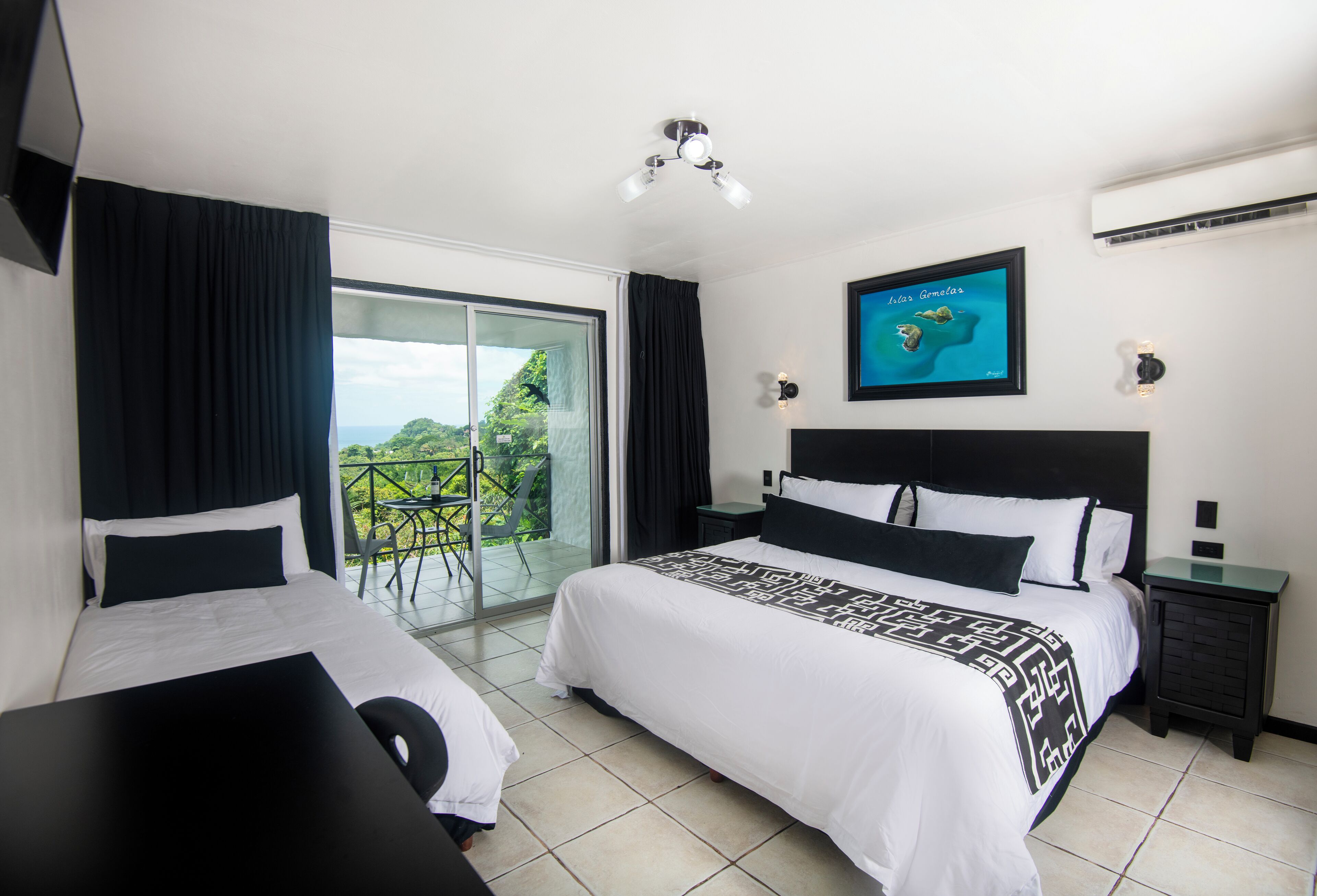 A hotel room with two beds, a balcony with a view, a ceiling fan, and a framed picture on the wall.