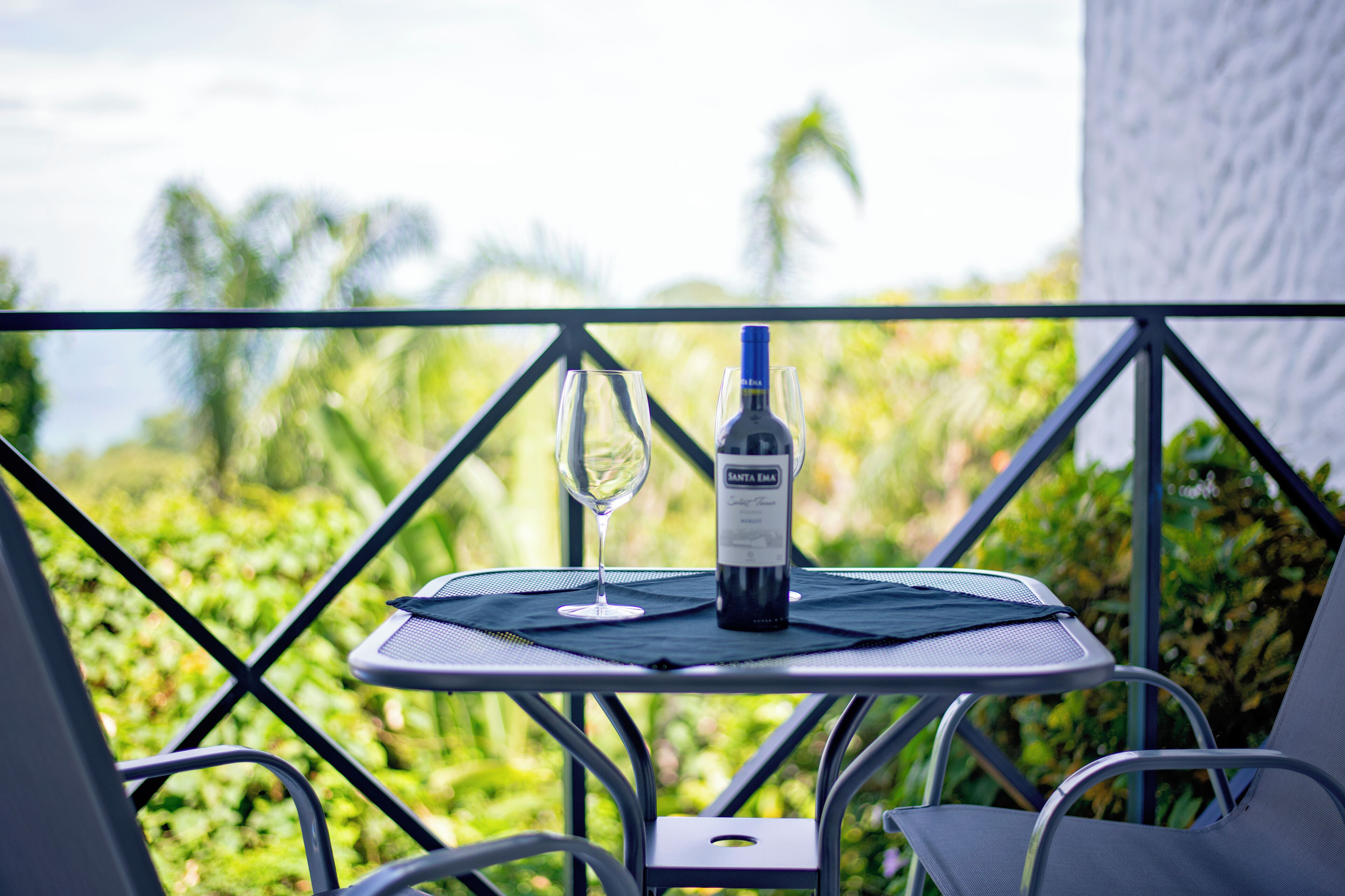 A table with a bottle of wine and a glass on a balcony with a view of greenery and a building in the background.