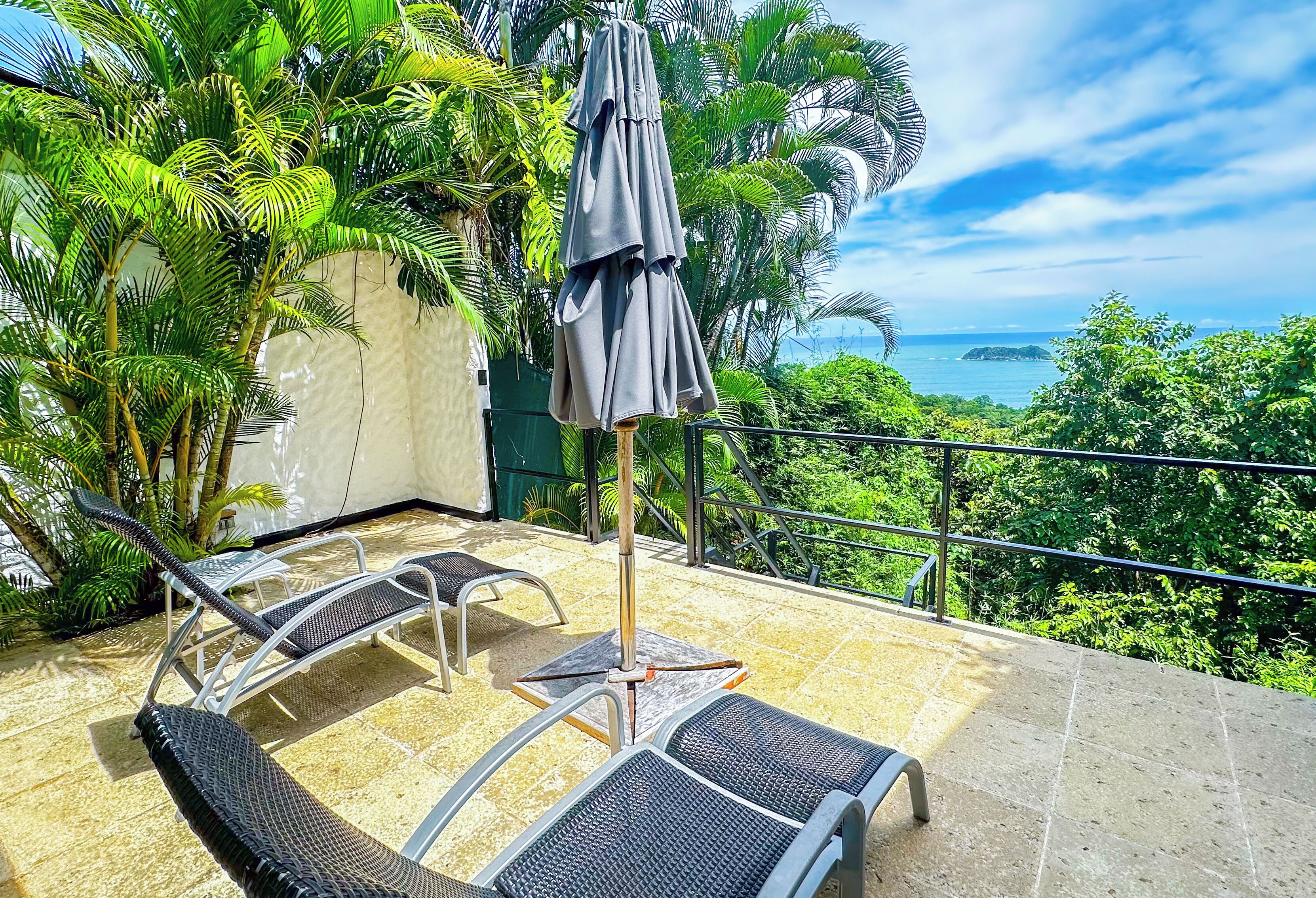 A balcony with two lounge chairs and a table, surrounded by lush greenery and a view of the sea.