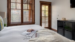 Een slaapkamer met een groot bed, houten zonwering, een tijdschrift, een hoed en een televisie aan de muur.