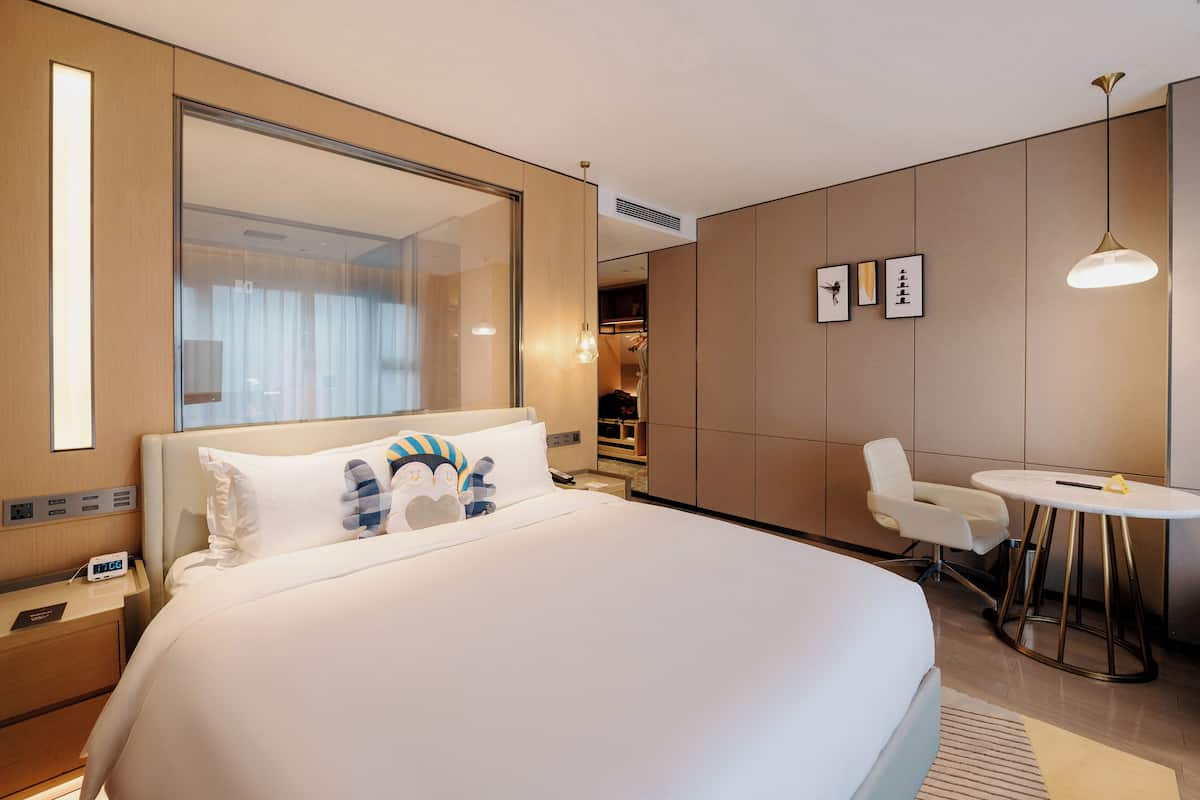 A modern hotel room with a large bed, a bedside table with a phone, a small round table with a chair, and a large mirror.