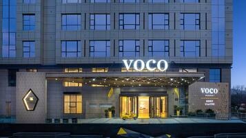 A modern hotel entrance with a prominent "VOCO" sign, a covered entrance area, and a yellow scooter parked outside.