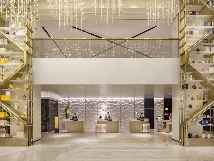 A modern hotel lobby with a central reception desk, two symmetrical staircases, and a decorative ceiling.