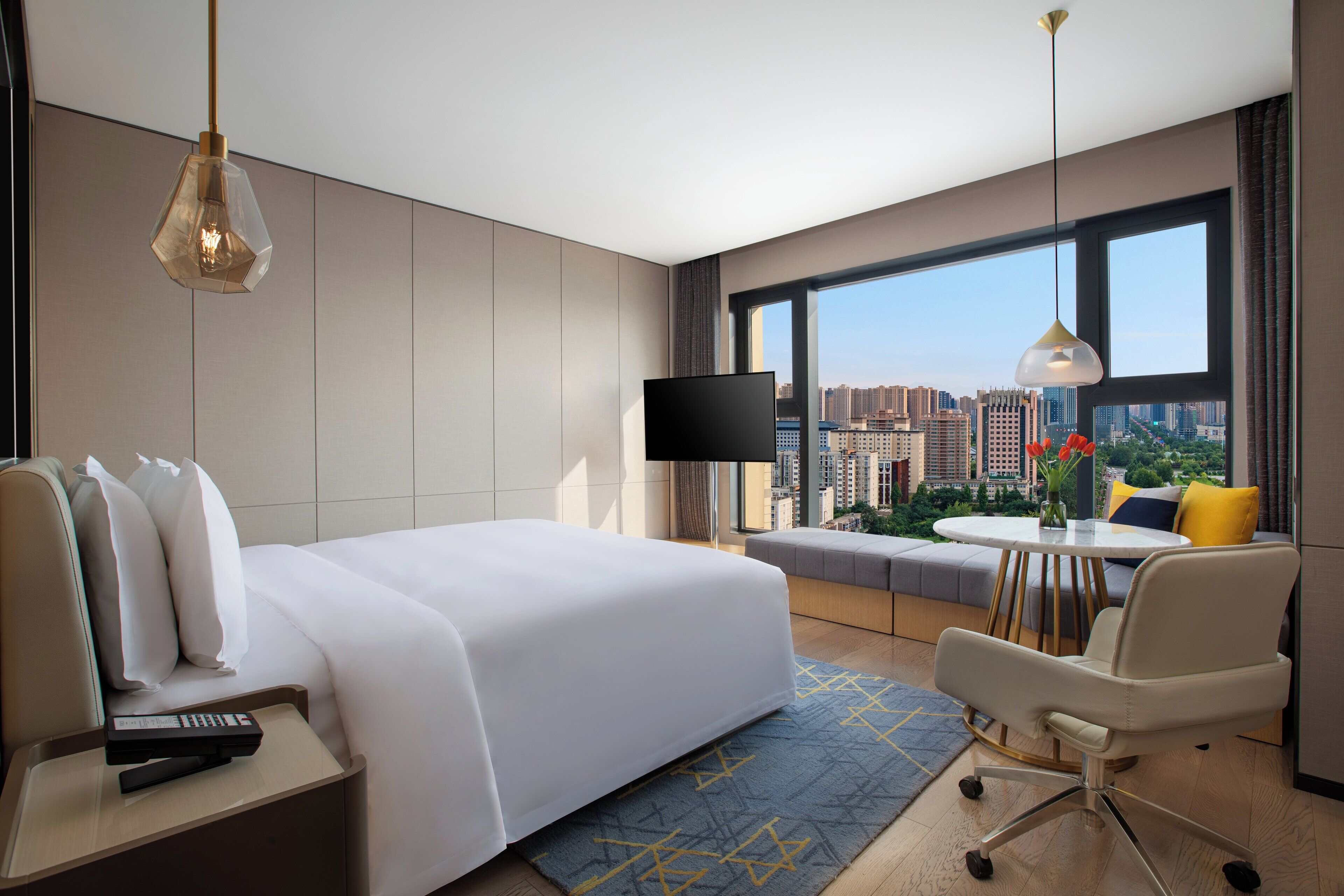 A modern hotel room with a large bed, a desk with a TV, a chair, and a view of the city through large windows.