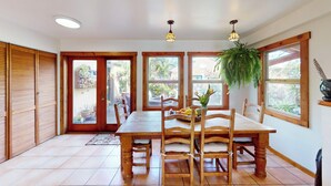 A dining area with a wooden table and chairs, large windows, and a hanging plant.