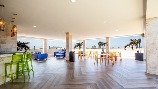 A spacious indoor area with a wooden floor, a bar area with green bar stools, a blue sofa, and a dining area with yellow chairs.