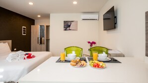 A hotel room with a neatly made bed, a breakfast table with fruit and muffins, and a TV mounted on the wall.