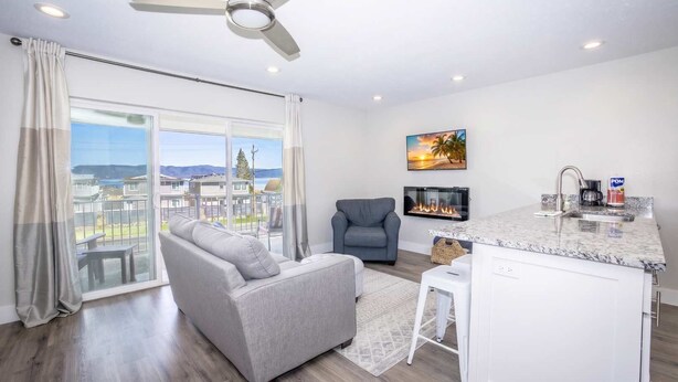 Een moderne woonkamer met een grijze bank, een grijze fauteuil, een flatscreen-televisie, een open haard en een keuken met een marmeren aanrechtblad en spoelbak.