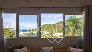 A room with a view of a beach and palm trees through large windows. There are two sofas with pillows labeled "ORIGINAL LIMITED". - Exceptional Villa Anse Marcel (Anse Marcel)