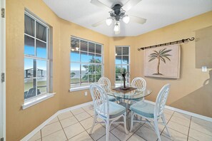 A dining area with a glass table, white chairs, a ceiling fan, and large windows with a view.