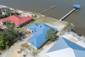 An aerial view of a property with two buildings, one with a red roof and another with a blue roof, adjacent to a body of water with a dock extending into it.