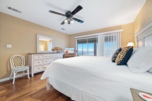 A bedroom with a large bed, a ceiling fan, a dresser, a chair, and a view of the ocean through a sliding door.