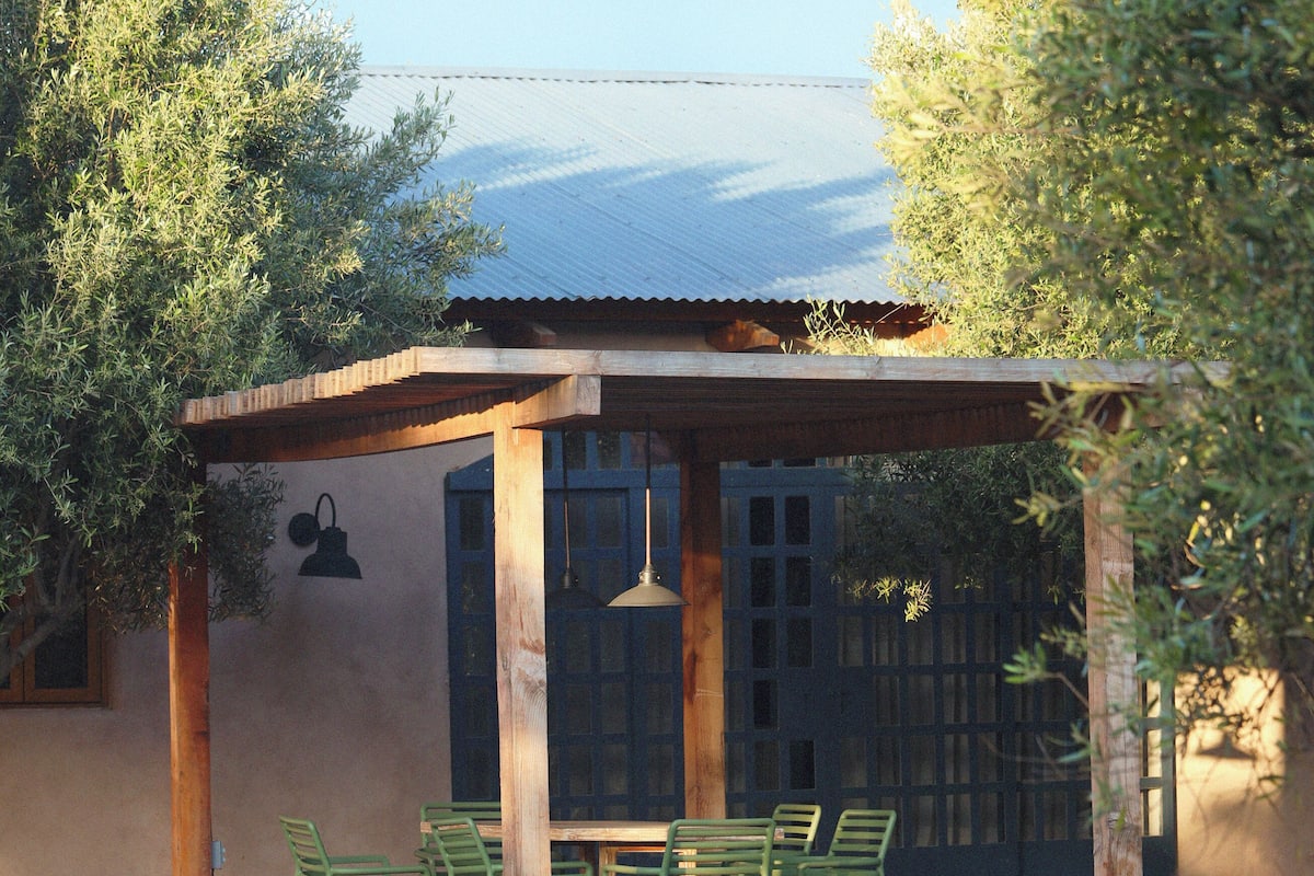 A covered outdoor seating area with wooden chairs and a table, surrounded by a gravel ground and a building with a corrugated metal roof.