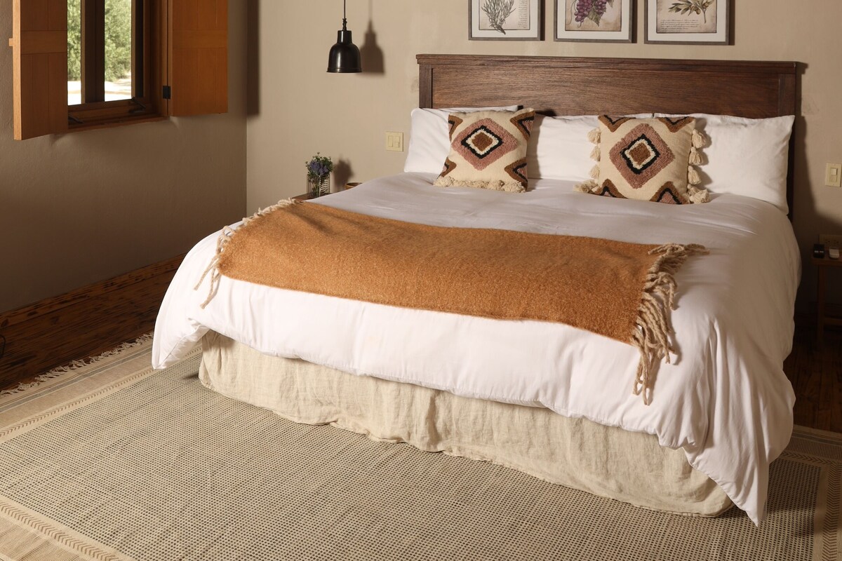 A bedroom with a wooden ceiling, a large bed with white bedding and patterned pillows, a window with open shutters, and framed botanical prints on the wall.