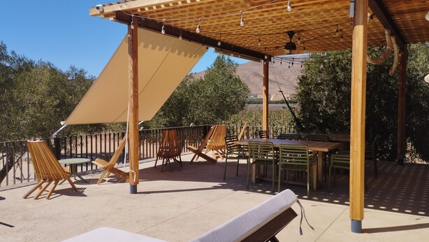 An outdoor patio with wooden furniture, a pergola, and a view of a vineyard. - Alma Liebre Viñedos del Valle (Valle de Guadalupe)