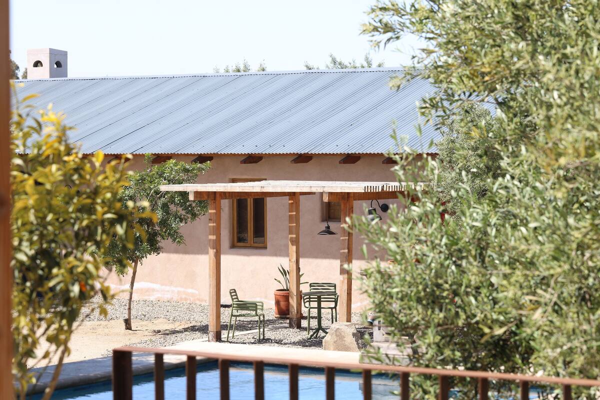 A patio with two chairs and a table, a small building with a metal roof, and a pool in the foreground.