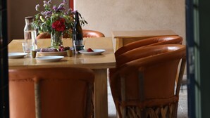A dining area with a table set for two, a wicker pendant light, framed pictures on the wall, and a vase with flowers on the table. - Alma Liebre Viñedos del Valle (Valle de Guadalupe)