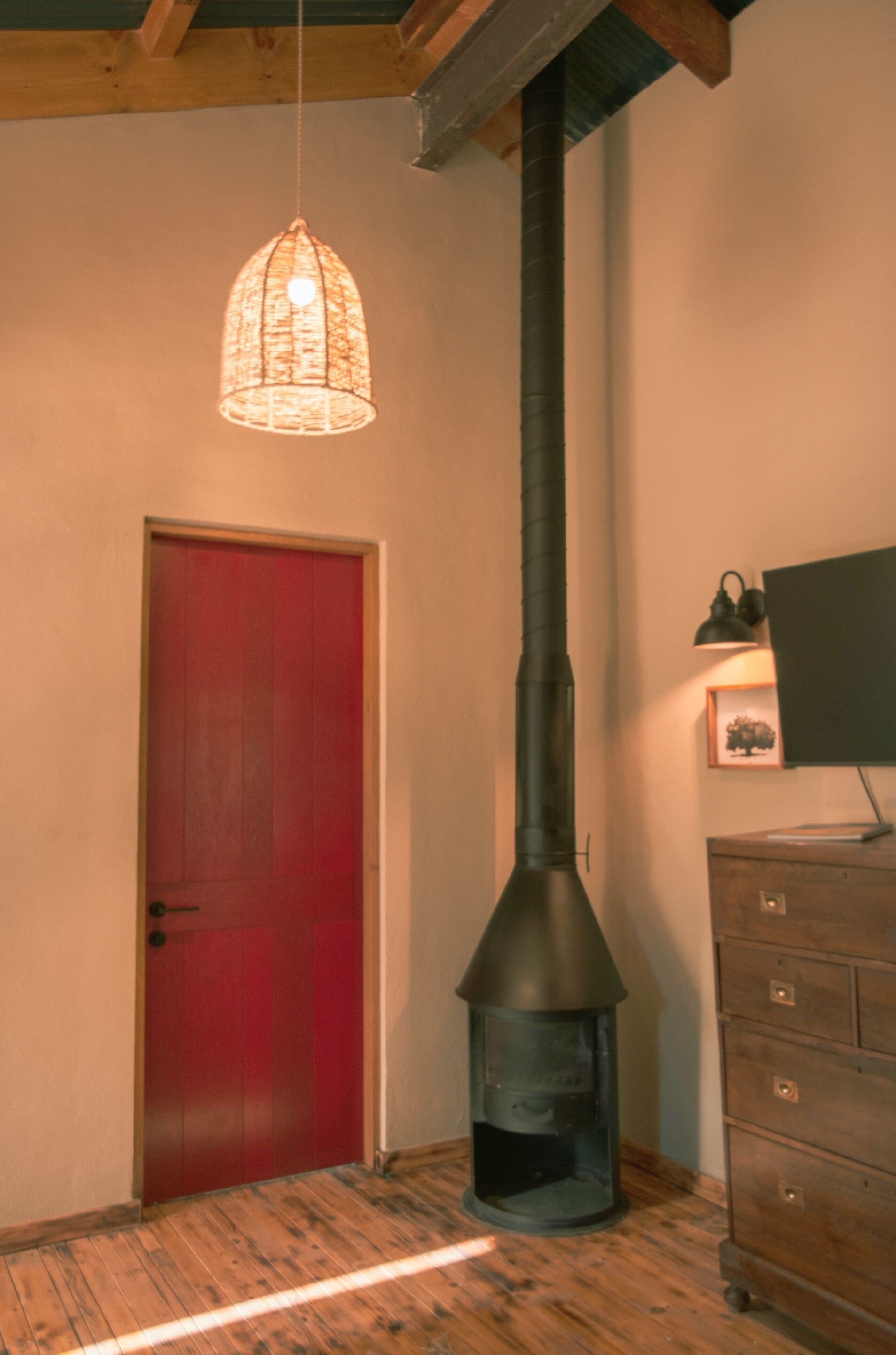 A room with a red door, a wooden floor, a black chimney, and a hanging light fixture.