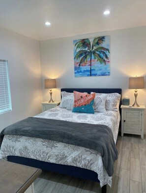 A bedroom with a bed, two bedside tables, a nightstand, a painting, and a window with blinds.