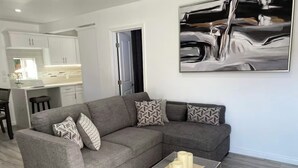 Ein modernes Wohnzimmer mit einem grauen Ecksofa, gemusterten Kissen, einem transparenten Glastisch und einem großen abstrakten Gemälde an der Wand.