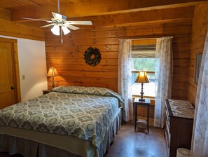 A cozy cabin bedroom with a bed, a ceiling fan, a window with curtains, and a small table with a lamp.