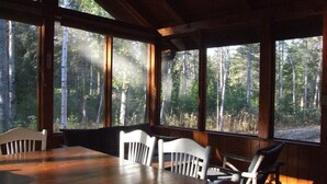 A wooden dining area with a large table and white chairs, large windows with a forest view, and a sloped ceiling.