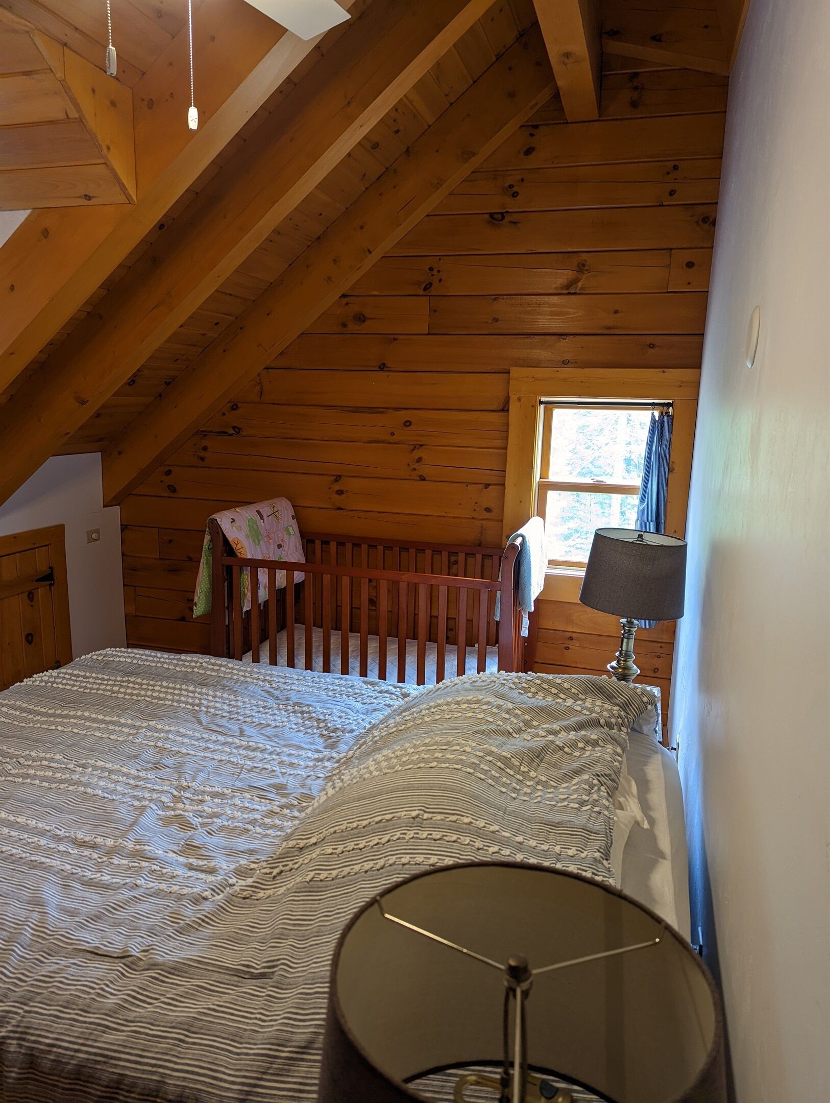 Un dormitorio con techo de madera, una cama con ropa de cama estampada, una cuna, una lámpara y una ventana con cortinas.