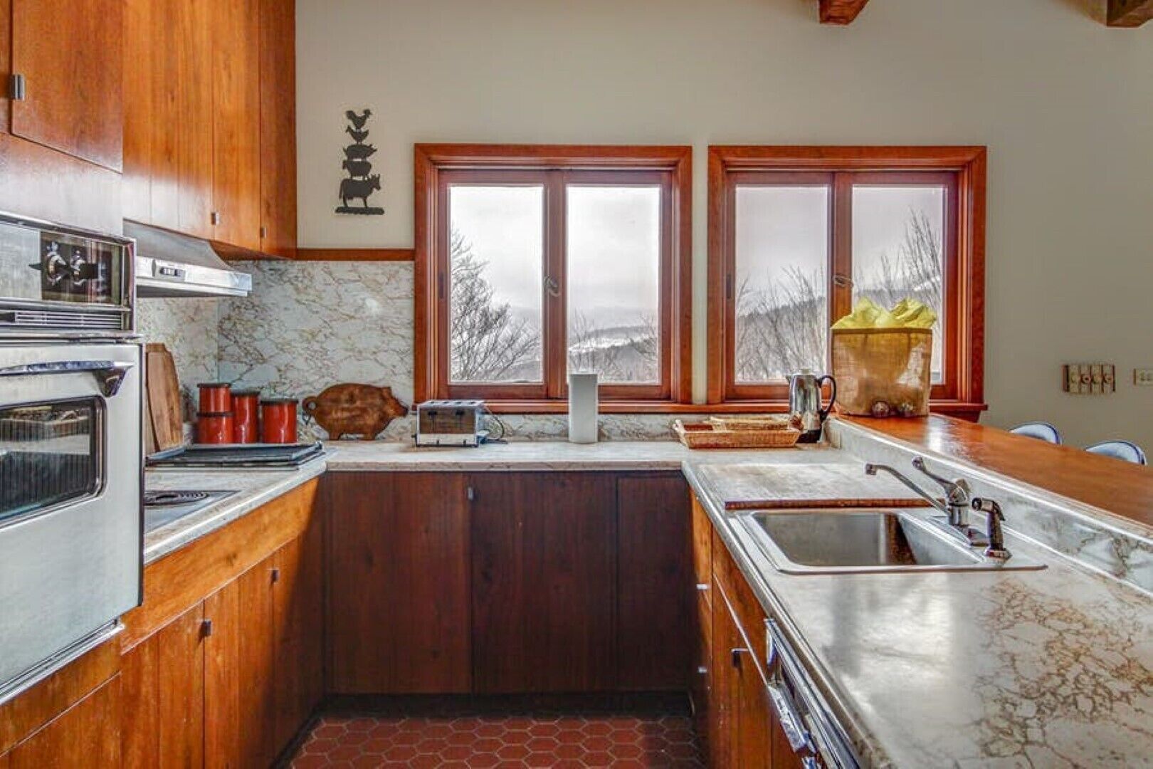 Una cocina con armarios de madera, estufa, fregadero y dos ventanas con vista a árboles.