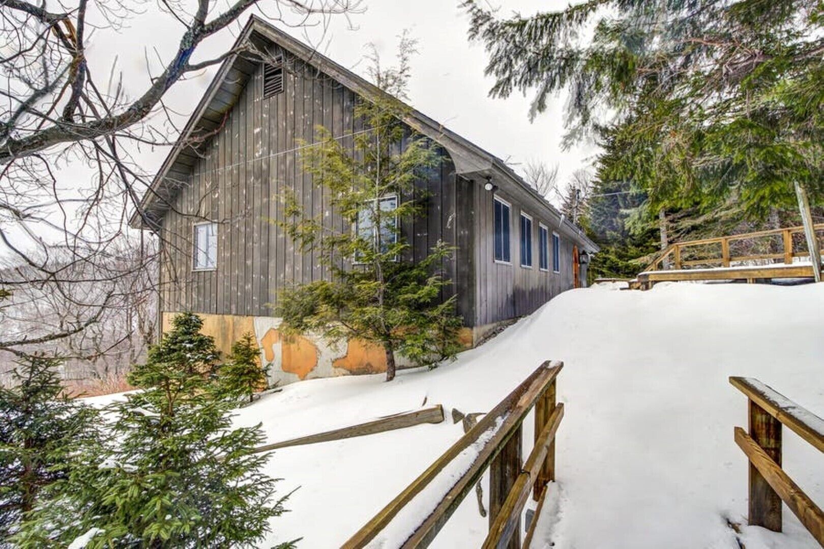 A rustic wooden cabin with a sloped roof, surrounded by snow-covered ground and trees.