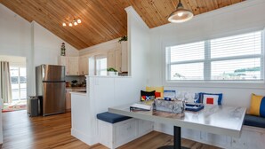 A modern kitchen with wooden ceiling, white cabinets, a stainless steel refrigerator, and a dining area with a bench and table.