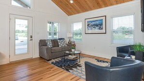 A spacious living room with a wooden ceiling, a gray sofa with patterned cushions, a coffee table, and a large window with blinds.