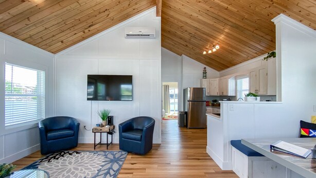 A modern interior with a wooden ceiling, a flat-screen TV mounted on the wall, a kitchen area with white cabinets, and a blue chair in the living space.