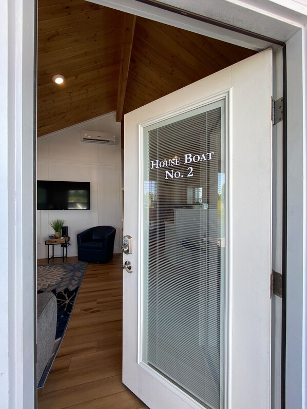 A modern interior with a wooden ceiling, a white door labeled "House Boat No. 2", a flat-screen TV, a small table with a plant, and a blue chair.