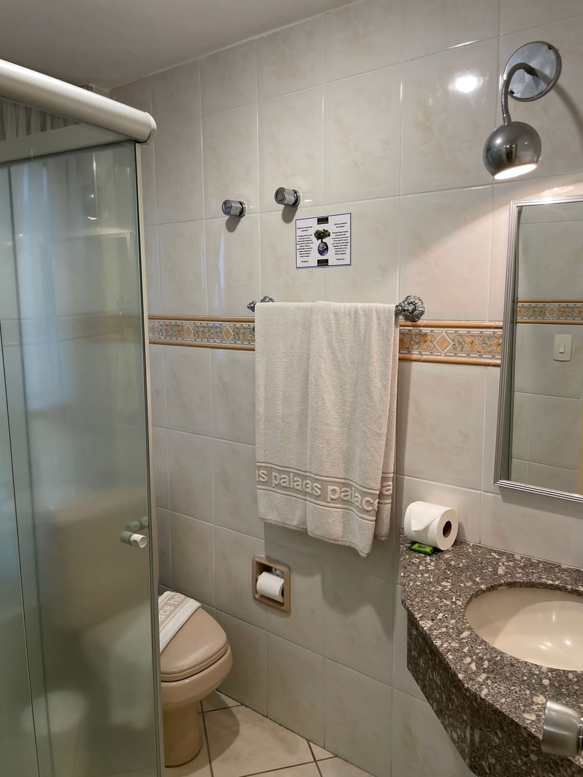 Un baño con cabina de ducha de vidrio, inodoro, lavamanos con encimera de mármol, toalla en un perchero y espejo instalado en la pared.