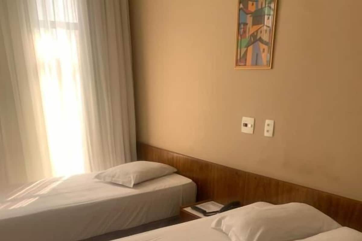 Une chambre d’hôtel avec deux lits simples, une tête de lit en bois, un tableau au mur et une fenêtre avec des rideaux.