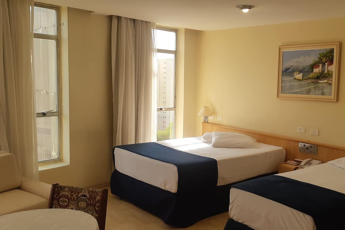 Une chambre d’hôtel avec deux lits simples, un canapé, un tableau au mur et une fenêtre avec des rideaux.
