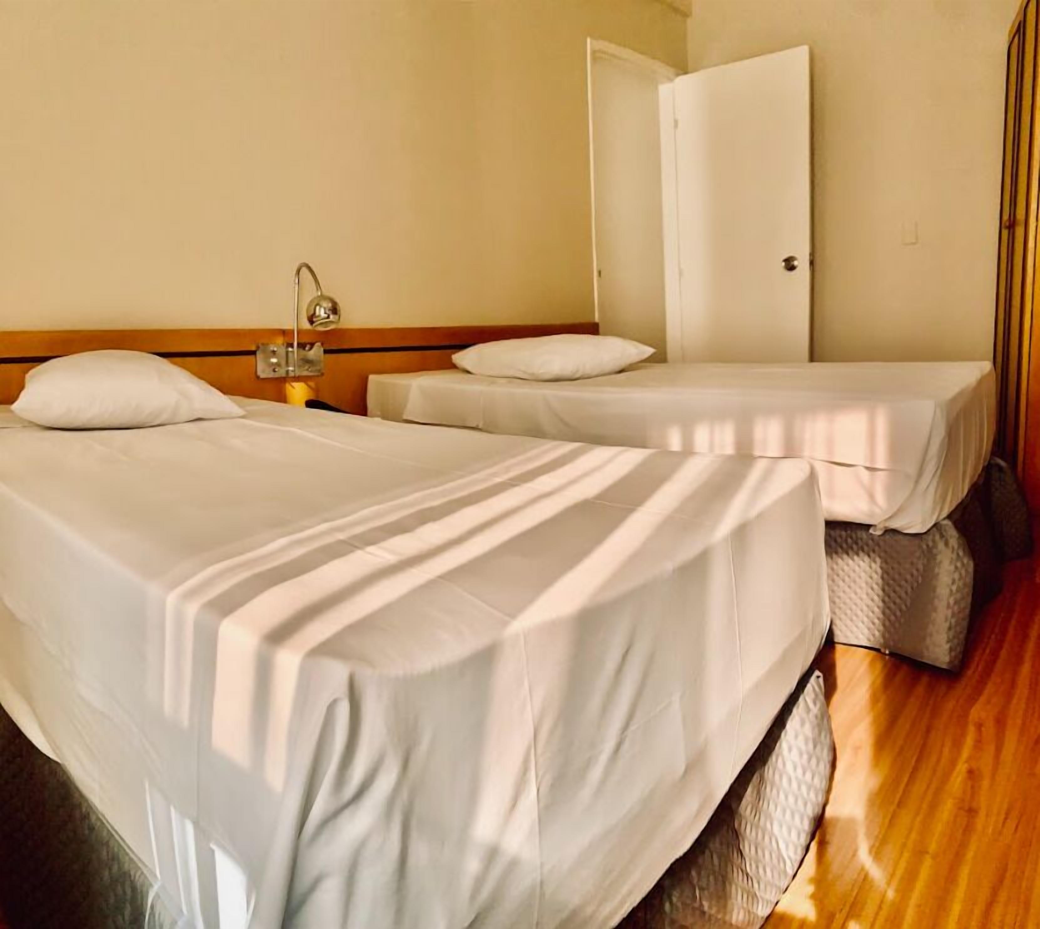 Quarto de hotel com duas camas de solteiro, cabeceira de madeira, mesa de cabeceira com abajur e uma porta à direita.