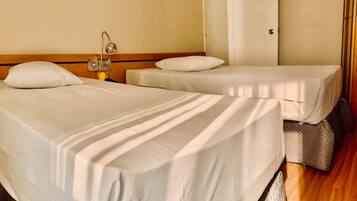 Quarto de hotel com duas camas de solteiro, cabeceira de madeira, mesa de cabeceira com abajur e uma porta à direita.