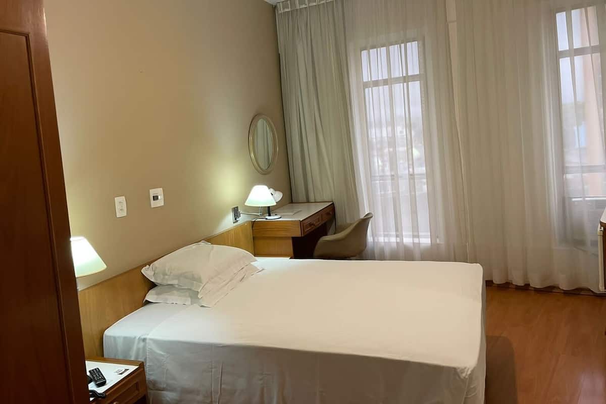 Une chambre d’hôtel avec un lit, un bureau, une chaise, une lampe et une fenêtre avec des rideaux.
