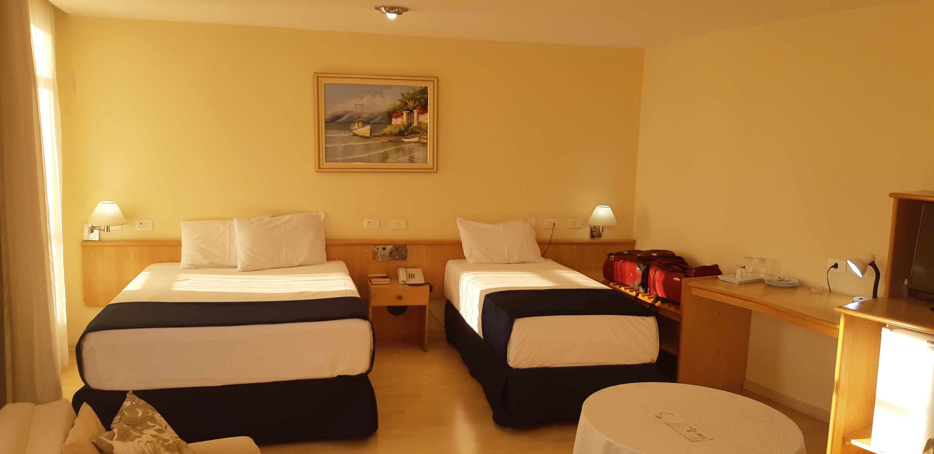 A hotel room with two beds, a desk, a chair, a small table, a painting on the wall, and a lamp.