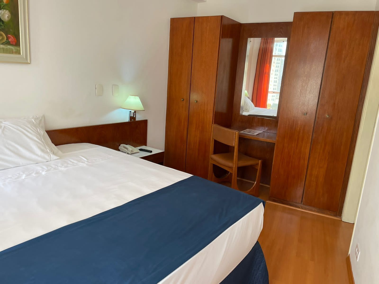 Quarto de hotel com cama, escrivaninha, cadeira, luminária e janela com cortinas.