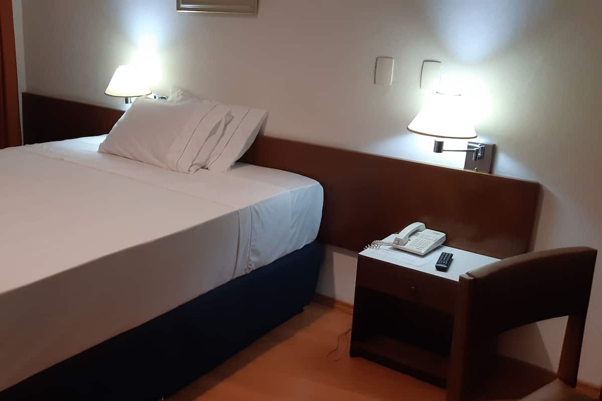 Une chambre d’hôtel avec un lit soigneusement fait, une table de chevet avec un téléphone et des documents, et deux lampes fixées au mur.