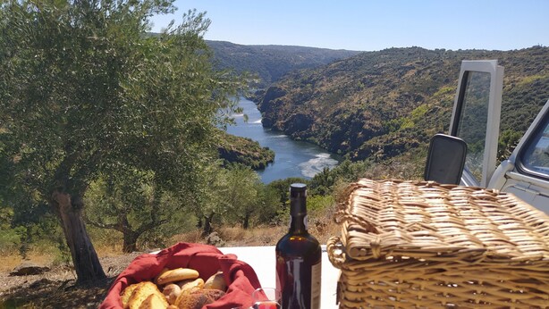Ein Tisch mit einem Brotkorb, einer Weinflasche und Gläsern, mit Blick auf ein malerisches Tal und einen Fluss.
