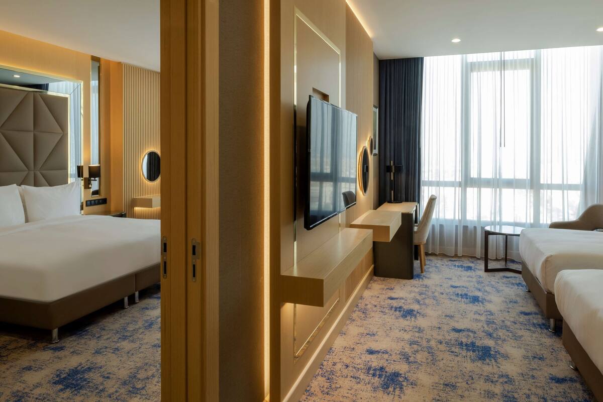 A hotel room with a large bed, a desk with a chair, a TV mounted on the wall, and a large window with curtains.