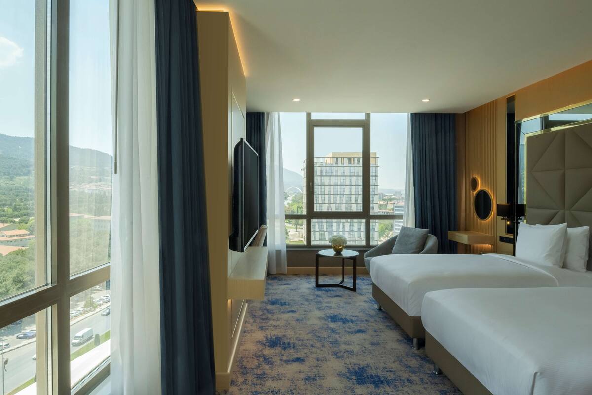 A hotel room with two beds, a TV, a small table with a lamp, and a view of a cityscape.