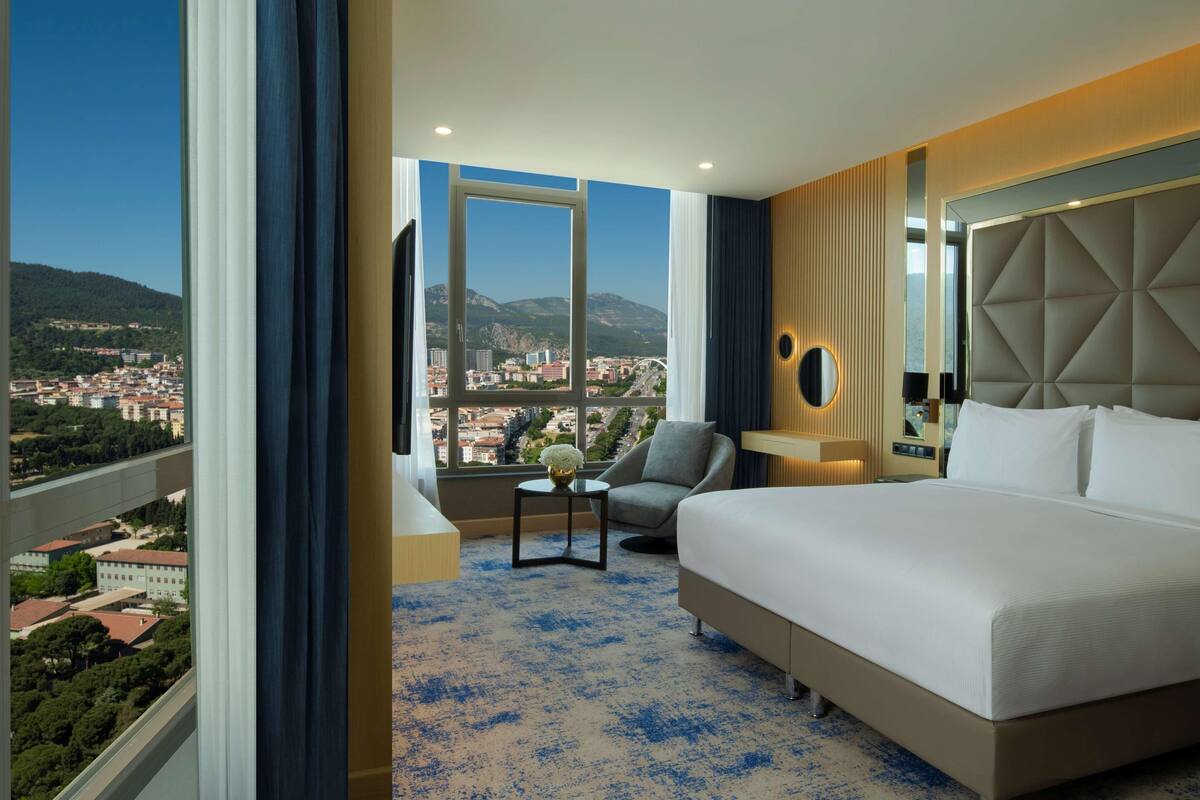 A hotel room with a large bed, a chair, a small table, and a view of a cityscape through large windows.