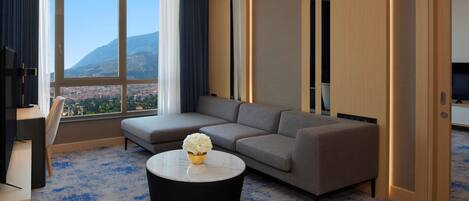A modern hotel room with a large window offering a cityscape view, a grey sofa, a black coffee table, and a TV on a wooden stand.